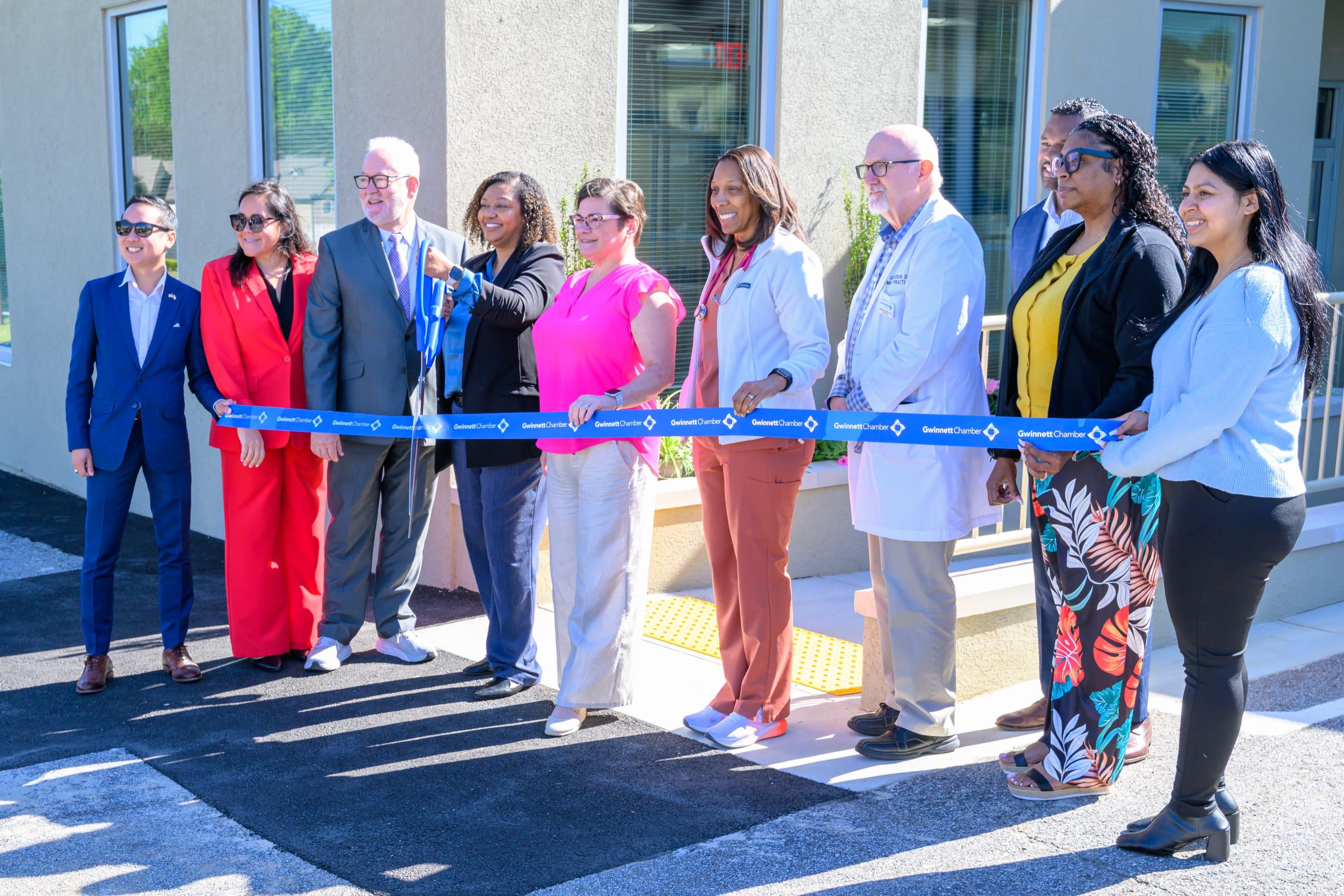 A group of people standing in front of a health clinic holding a long blue ribbon with Good Samaritan Health Center written on it. They are preparing to cut the ribbon to open the new center.