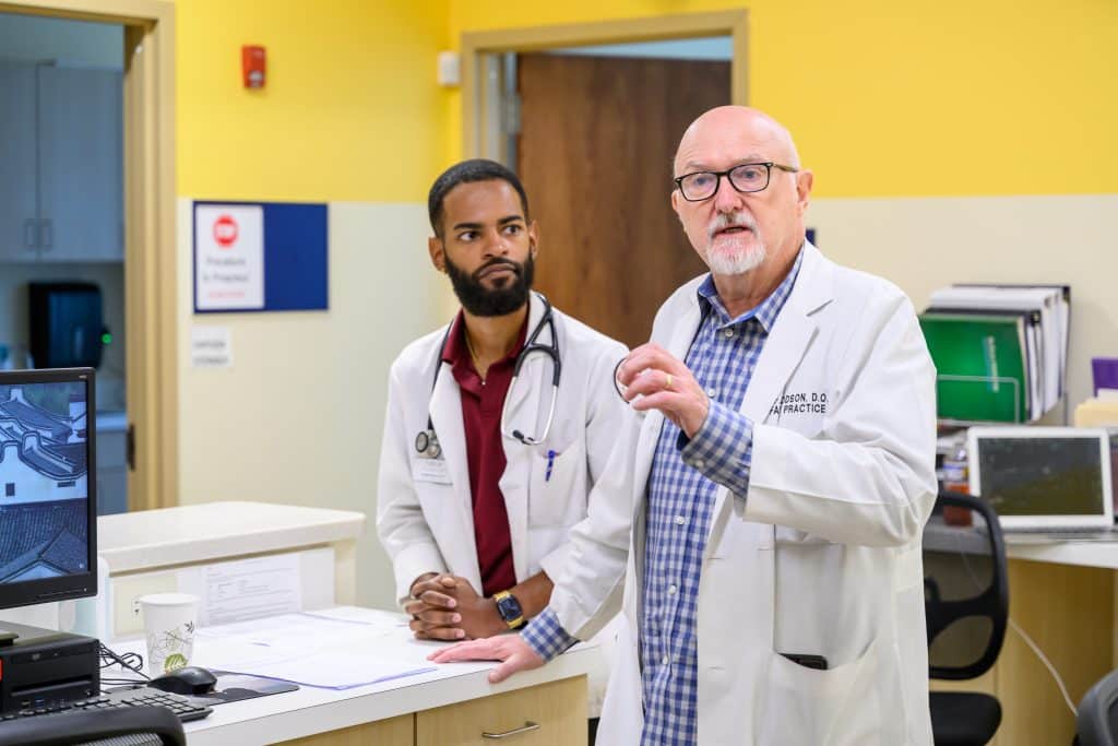 A young Black physician and an older white male physician standing at the nurse's station in a healthcare center. They are talking to a group of people (not shown in the photo) that are taking a tour of the facility. There are computers on the desk along with papers and other items. The wall behind them is bright yellow and white.