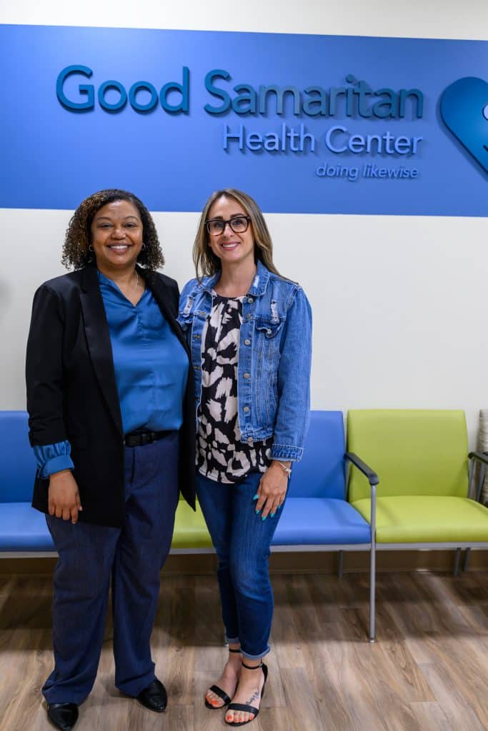 A Black woman and white woman standing together in the waiting room of a community health center. They're both smiling. The blue and white wall behind them has the words "Good Samaritan Health Center doing likewise" and a logo.