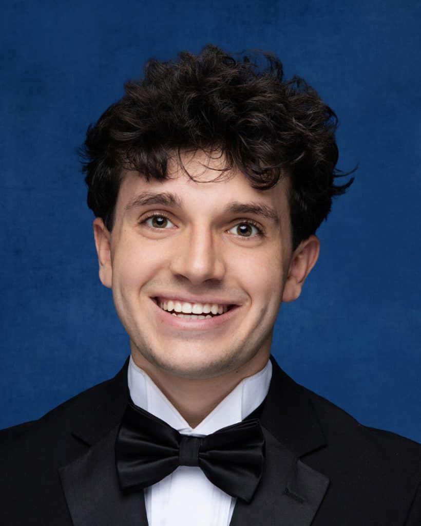 High School senior photo of a young white man with brown curly hair against a blue background. He's wearing a tux with bowtie and white shirt and smiling.