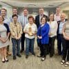 A group of people standing together for an award presentation in an office. A small elderly woman with short brown hair stands in the center holding an award for sustainability