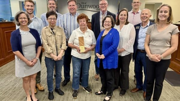 A group of people standing together for an award presentation in an office. A small elderly woman with short brown hair stands in the center holding an award for sustainability