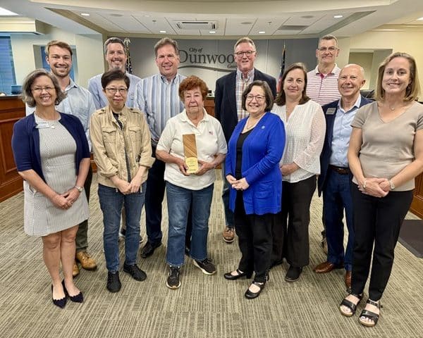 A group of people standing together for an award presentation in an office. A small elderly woman with short brown hair stands in the center holding an award for sustainability