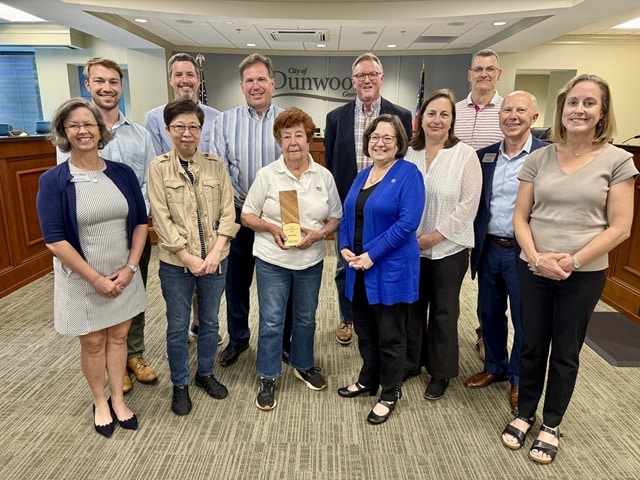 A group of people standing together for an award presentation in an office. A small elderly woman with short brown hair stands in the center holding an award for sustainability