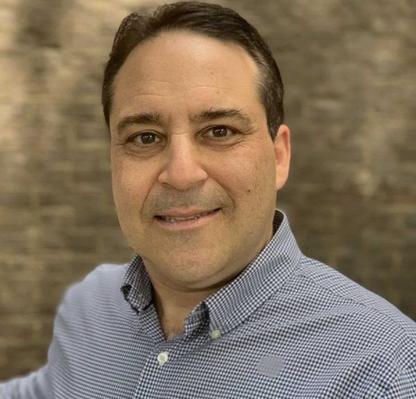 A middle-aged man with short dark hair, wearing a blue button-up shirt with collar. He's looking at the camera and smiling, standing in front of a brown and cream-colored brick background.