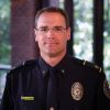 A white male police chief wearing uniform and badge and glasses. He's standing in the police department with windows behind him.