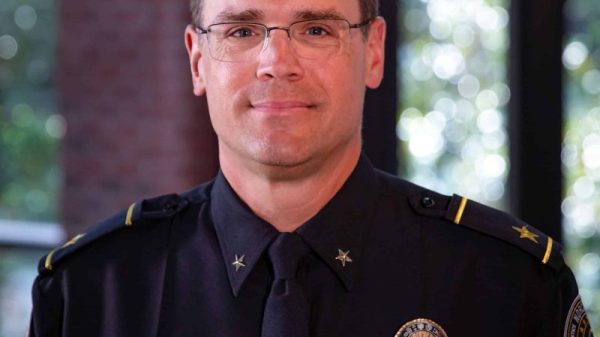 A white male police chief wearing uniform and badge and glasses. He's standing in the police department with windows behind him.