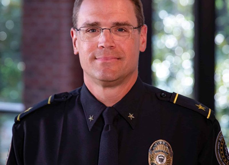 A white male police chief wearing uniform and badge and glasses. He's standing in the police department with windows behind him.