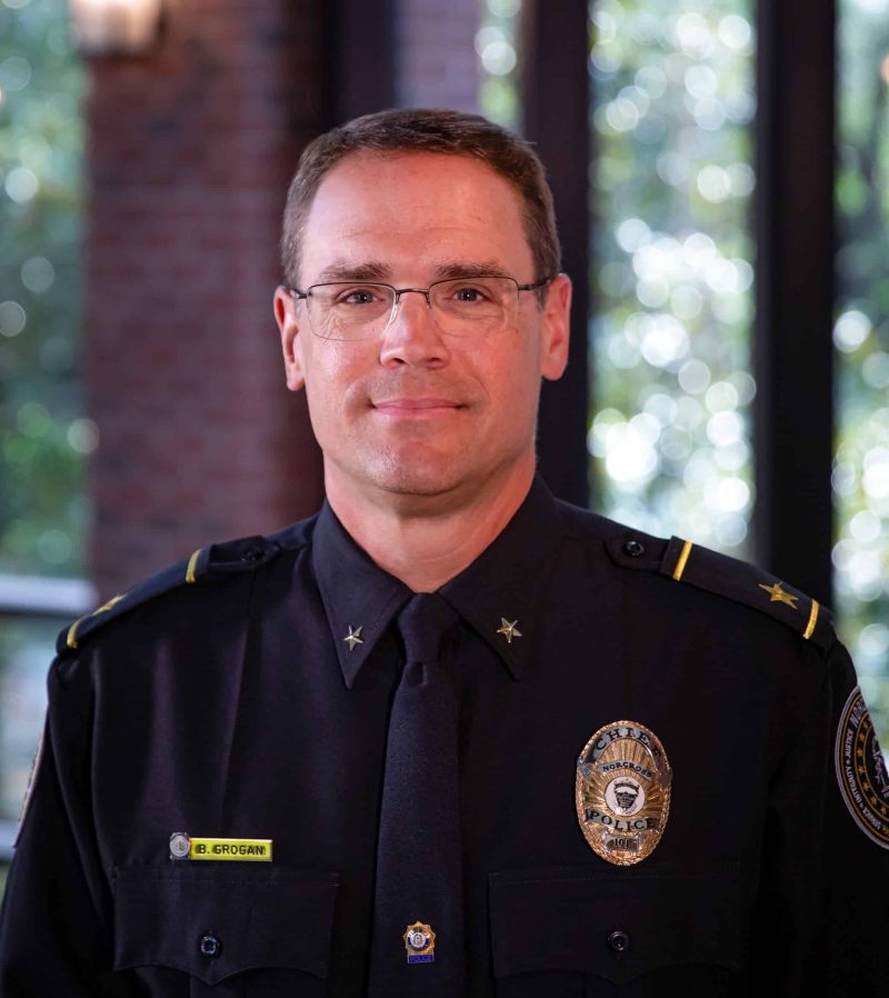 A white male police chief wearing uniform and badge and glasses. He's standing in the police department with windows behind him.