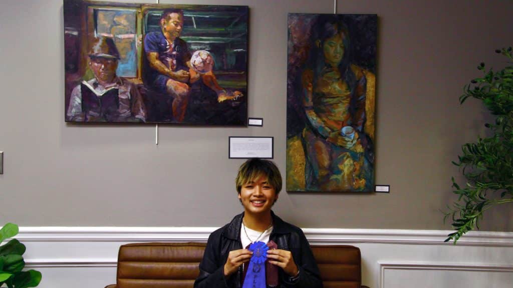 A high-school-aged artist holding a first place blue ribbon. He is sitting on a bench beneath his two winning paintings.
