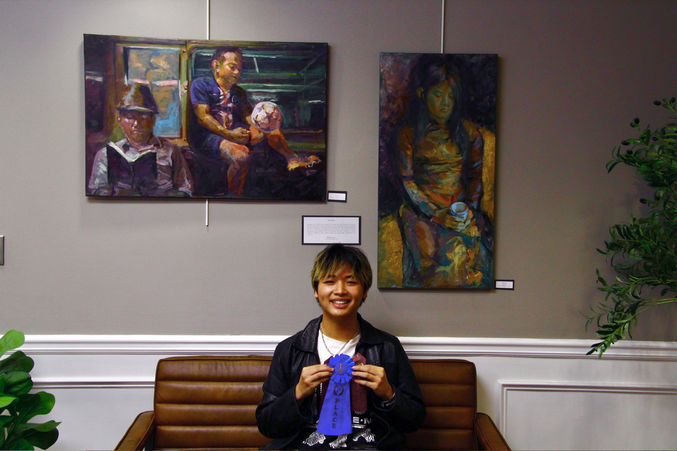 A high-school-aged artist holding a first place blue ribbon. He is sitting on a bench beneath his two winning paintings.