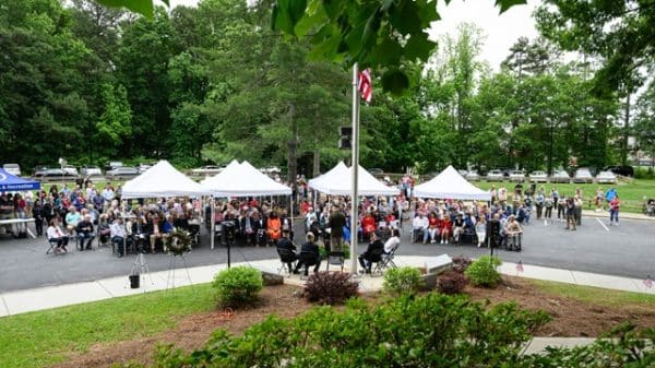 Several open-sided tents with white canvas tops set up in a parking lot. There's a grassy area and flagpole with an American flag in front and dozens of people sitting in folding chairs or standing in the crowd for a Memorial Day ceremony.