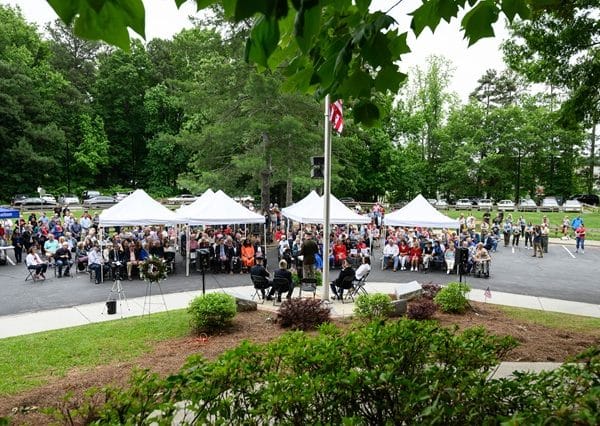 Several open-sided tents with white canvas tops set up in a parking lot. There's a grassy area and flagpole with an American flag in front and dozens of people sitting in folding chairs or standing in the crowd for a Memorial Day ceremony.