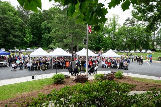 Several open-sided tents with white canvas tops set up in a parking lot. There's a grassy area and flagpole with an American flag in front and dozens of people sitting in folding chairs or standing in the crowd for a Memorial Day ceremony.