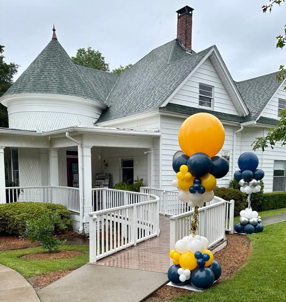 A white historic building with walkway. There are gold, black and white balloons at the entrance marking the opening of an art show.