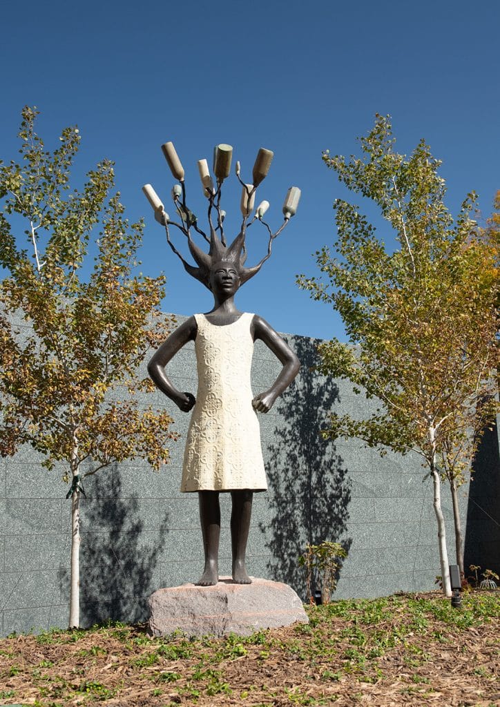 Little Big Sister outdoor sculpture by Alison Saar, made with cast stainless steel with patina. It shows a young Black girl with hands on hips, wearing a white dress and is set in a grassy area with small trees and a grey wall behind it.