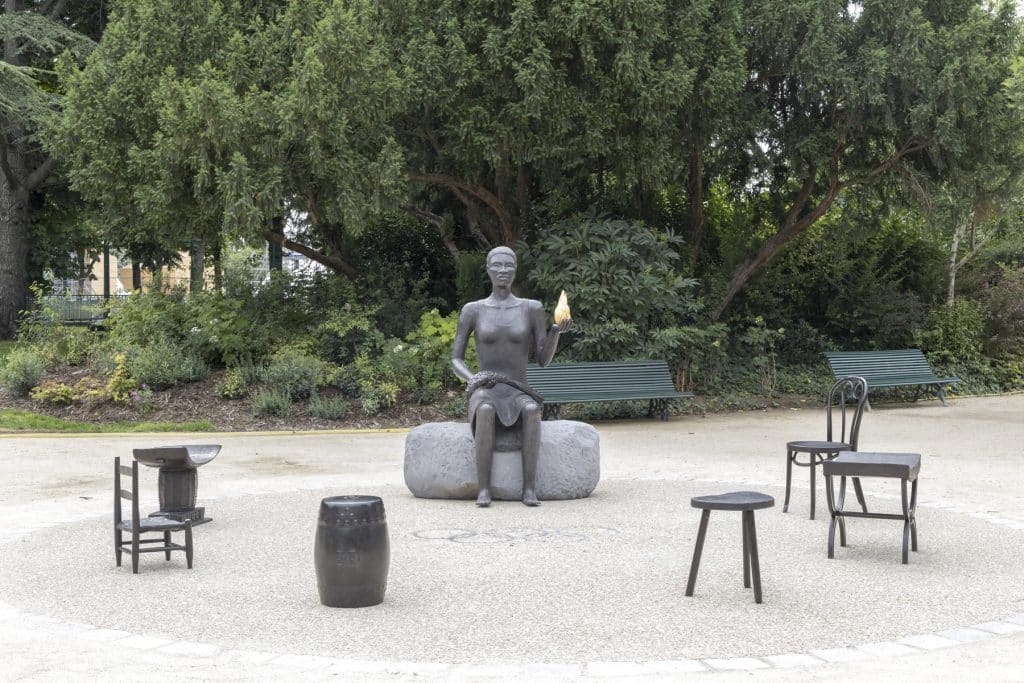 Cast bronze and stone outdoor sculpture, Salon, by Alison Saar in Paris. It shows a woman sitting on a large stone bench holding a golden object in her left hand, surrounded by smaller chairs and seats in a loose circle around her.