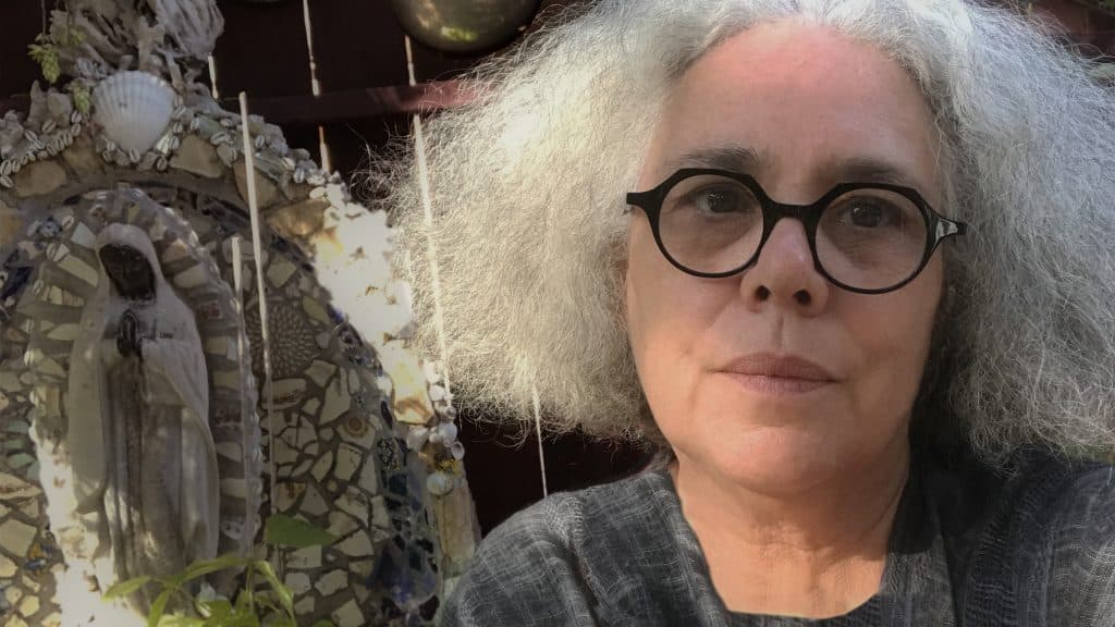Artist, Alison Saar. She has chin-length frizzy white hair parted in the middle. She's wearing round, black-frame glasses and a grey top. She's sitting in a garden with a small outdoor sculpture and lots of green plants behind her.