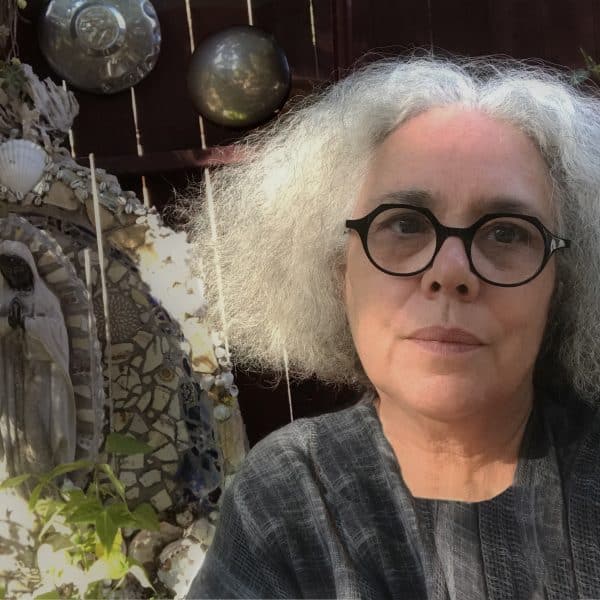 Artist, Alison Saar. She has chin-length frizzy white hair parted in the middle. She's wearing round, black-frame glasses and a grey top. She's sitting in a garden with a small outdoor sculpture and lots of green plants behind her.