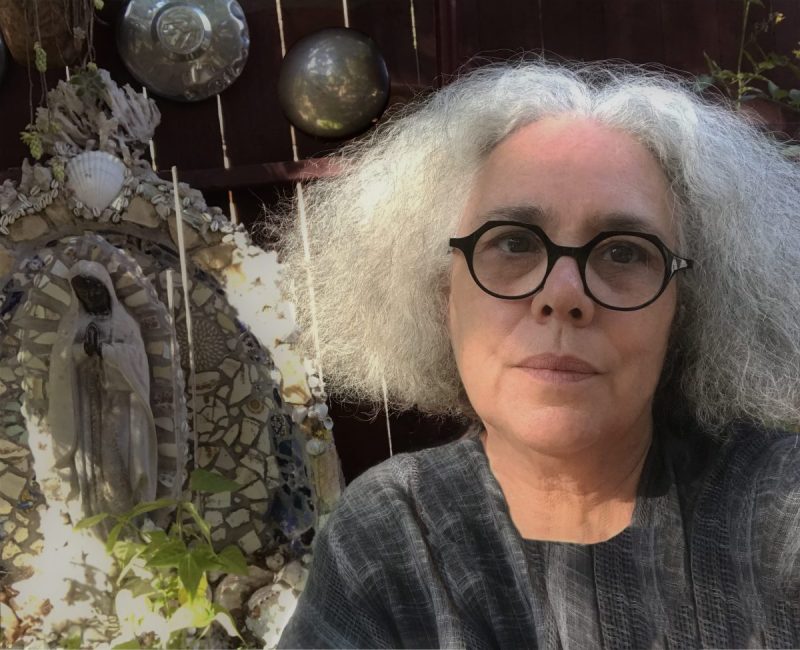 Artist, Alison Saar. She has chin-length frizzy white hair parted in the middle. She's wearing round, black-frame glasses and a grey top. She's sitting in a garden with a small outdoor sculpture and lots of green plants behind her.