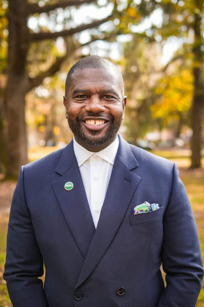 A Black man with short hair wearing a blue suit and white, button-up shirt. He's smiling and standing outside with trees and grass in the background.