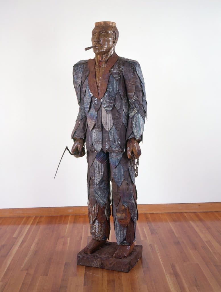 Alison Saar's Tobacco Demon sculpture, showing a man made up of tobacco leaves, holding a tool and smoking a cigarette, made of ceiling tin, found objects and shellac on wood
