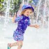 Young child wearing purple shorts, top and hat, splashing in a splash pad.