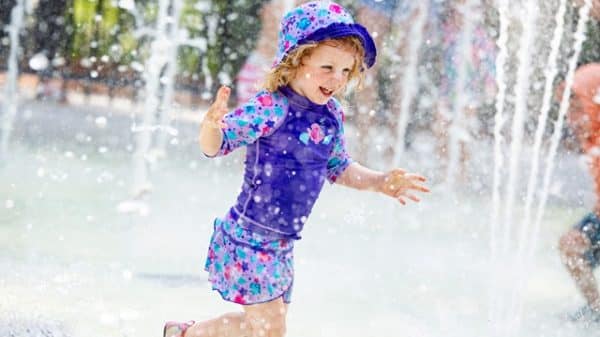 Young child wearing purple shorts, top and hat, splashing in a splash pad.