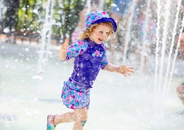 Young child wearing purple shorts, top and hat, splashing in a splash pad.