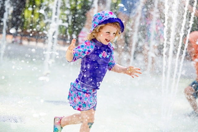 Young child wearing purple shorts, top and hat, splashing in a splash pad.