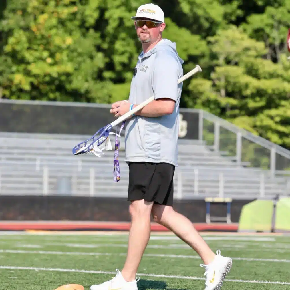 A white man in shorts and athletic clothes, wearing a light-colored cap and sunglasses. He's on a sports field holding a lacrosse stick under his left arm.