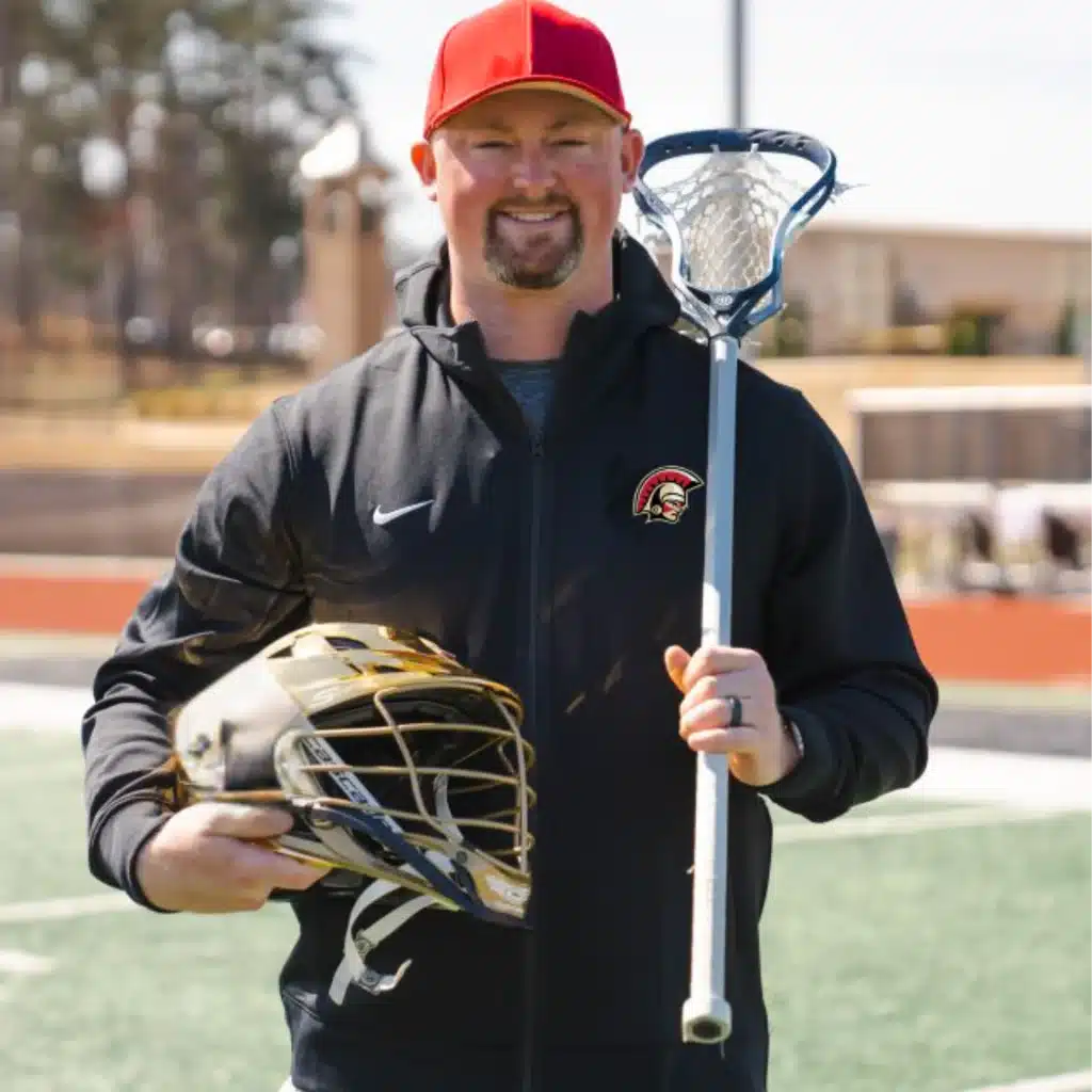A white man with short, dark facial hair, wearing a red cap and athletic clothes. He's standing on a high school sports field holding a lacrosse helmet and lacrosse stick.